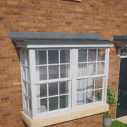 White bay window with grid panes, crafted with GRP Building Components, on a red brick house, next to a black door and green bushes.