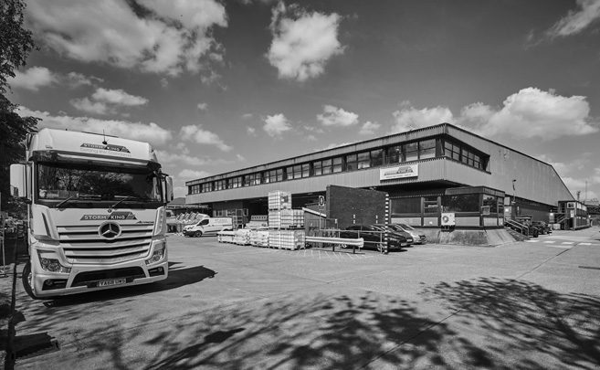 Black and white image of a warehouse with a lorry parked outside and pallets stacked nearby.