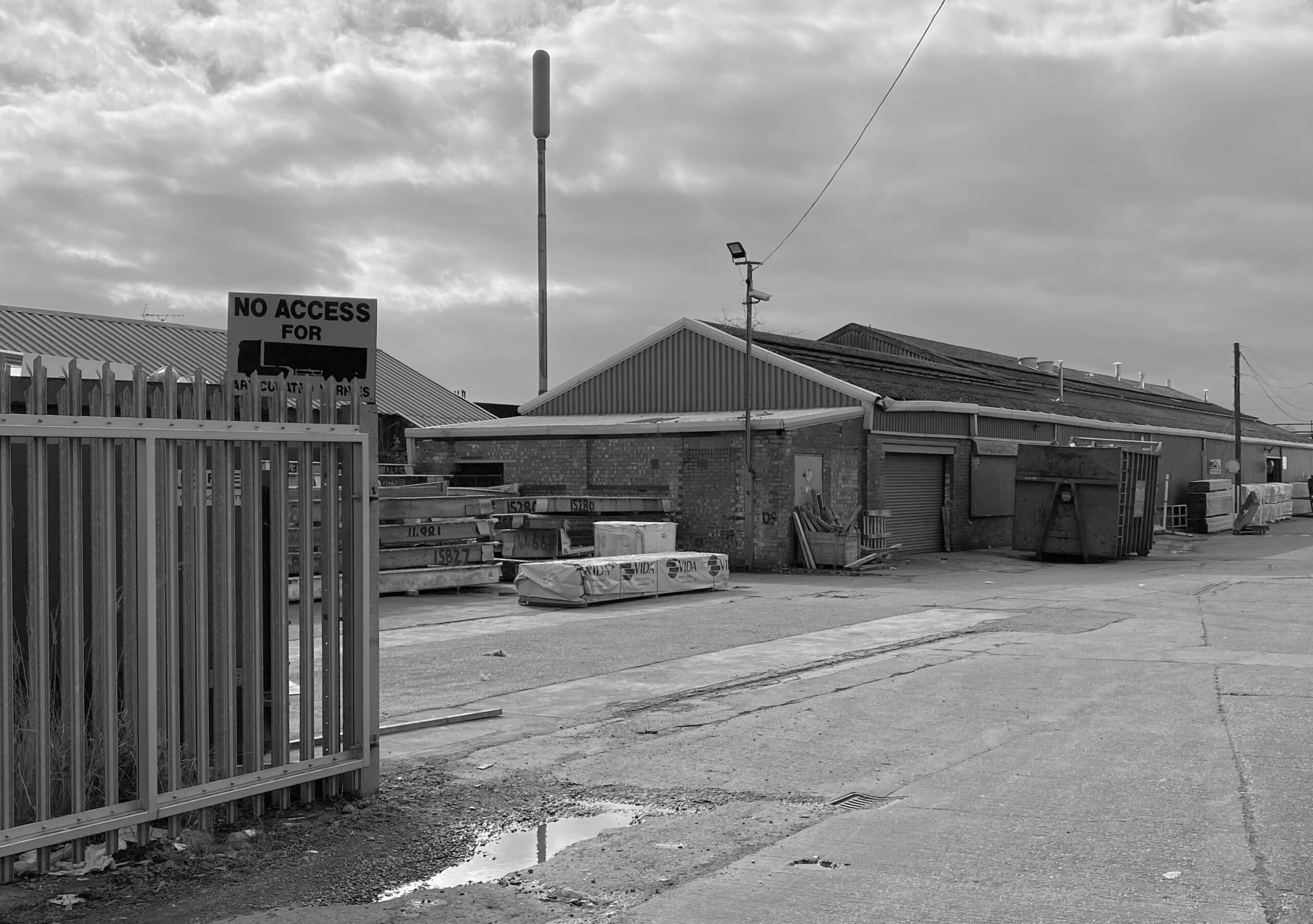 Black and white image of an industrial yard with a No Access For Vehicles sign on a fence.