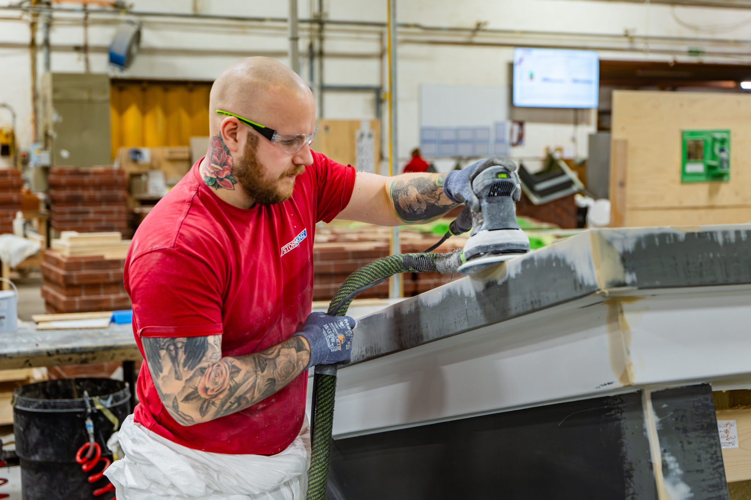 A man in safety gear sands a large GRP object in an industrial workshop.
