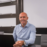 Bald man in a Stormking blue shirt sits at a desk with laptop and mobile, smiling at the camera.