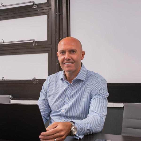 Bald man in a Stormking blue shirt sits at a desk with laptop and mobile, smiling at the camera.