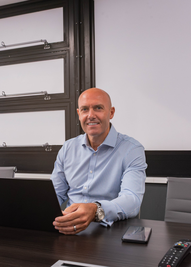Bald man in a Stormking blue shirt sits at a desk with laptop and mobile, smiling at the camera.
